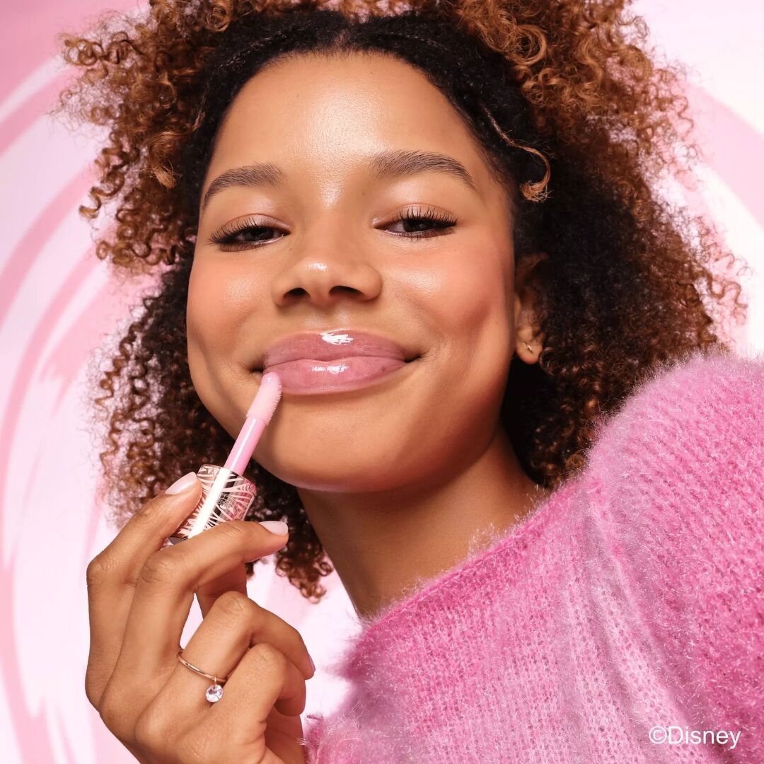 Woman applying Essence Disney plumping high shine lip gloss with a pink background